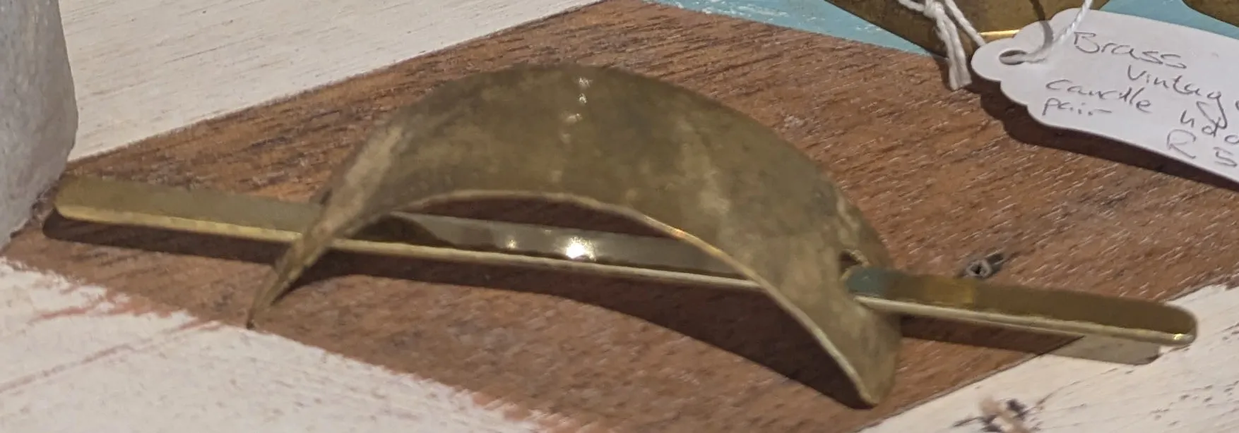 A crescent-shaped hammered brass hair clasp with a long brass pin lying on a painted wooden shelf
