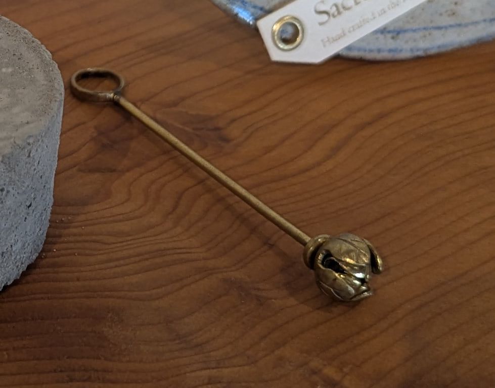 A slim brass stem with a loop at one end and a closed lotus bud at the other, lying on a wooden shelf