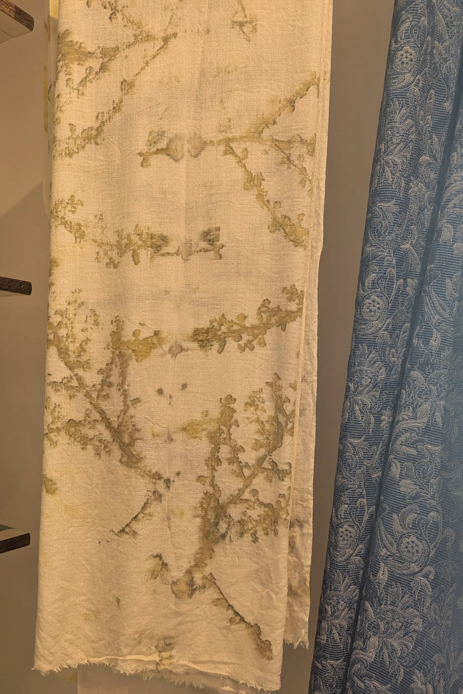 A cream cashmere scarf with olive leaf prints hanging beside a blue damask curtain