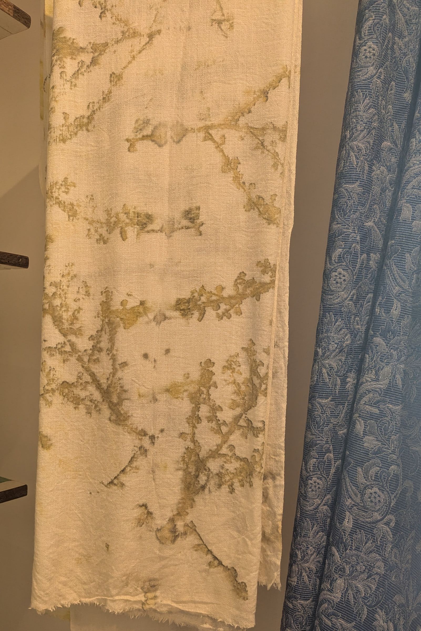 A cream cashmere scarf with olive leaf prints hanging beside a blue damask curtain