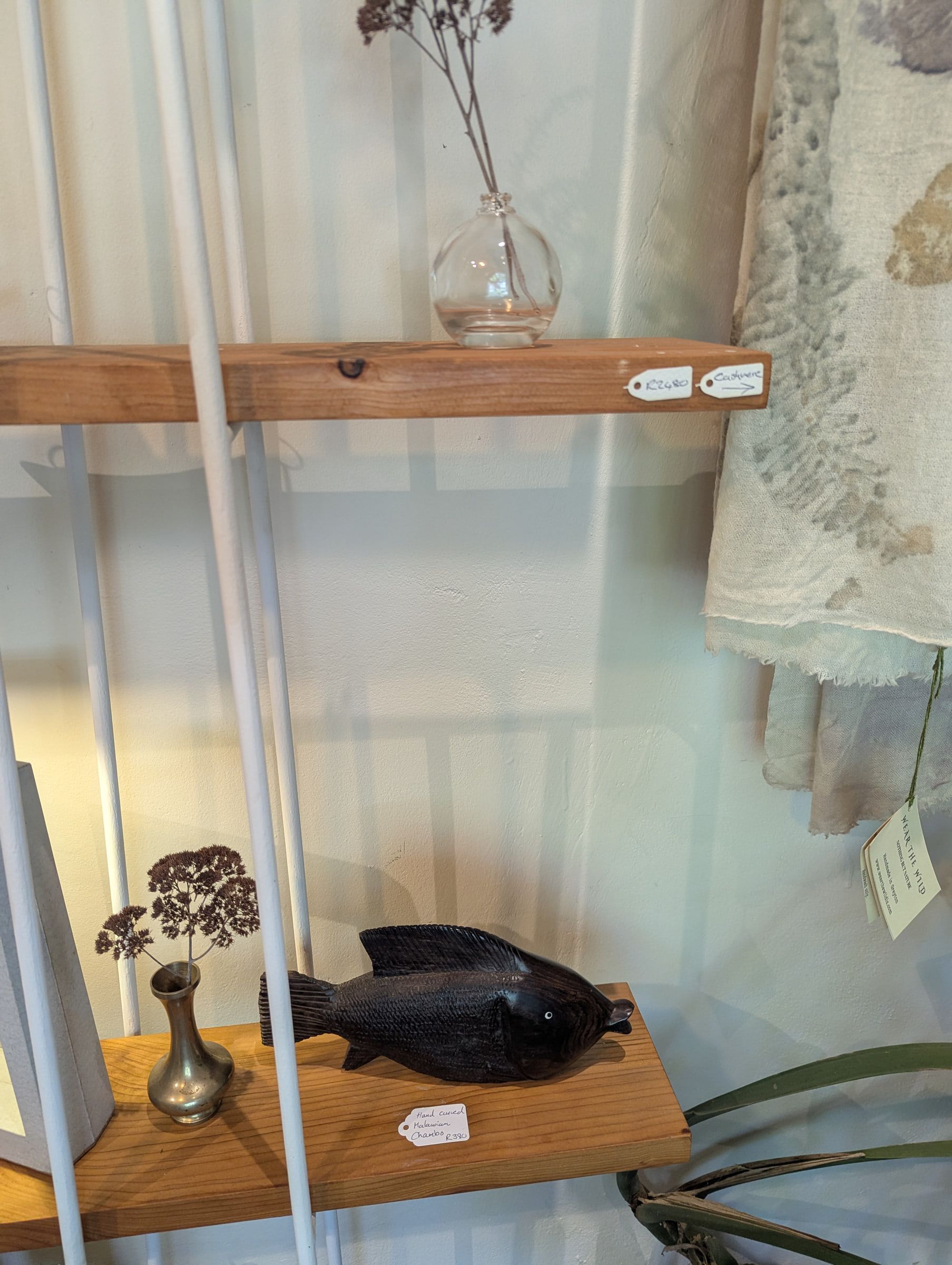 Morning light on a gallery shelf with a carved fish, a small brass vase and a leaf-printed scarf