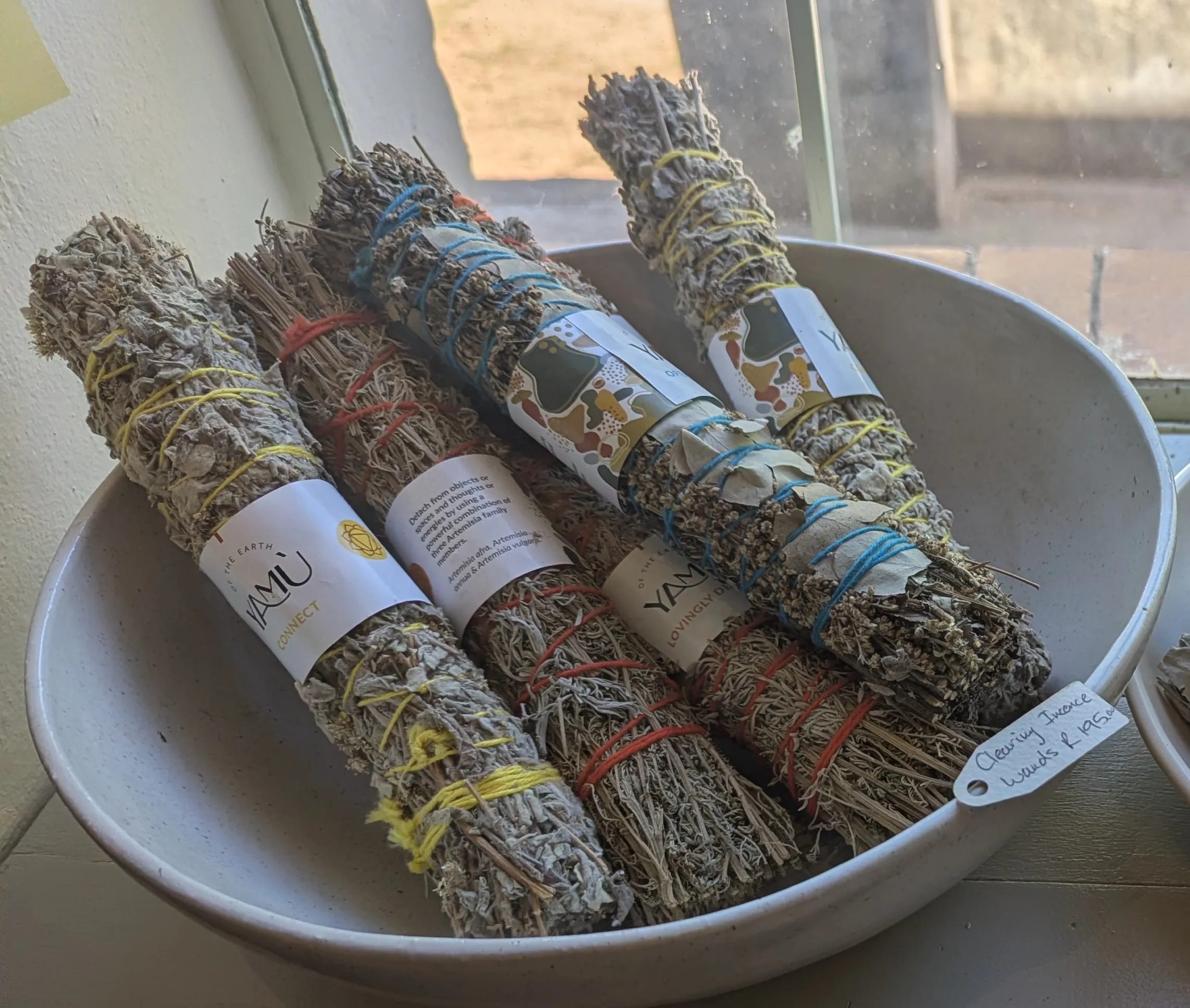 Hand-tied herb incense wands with coloured threads and YAMU labels in a white bowl on a sunny window sill