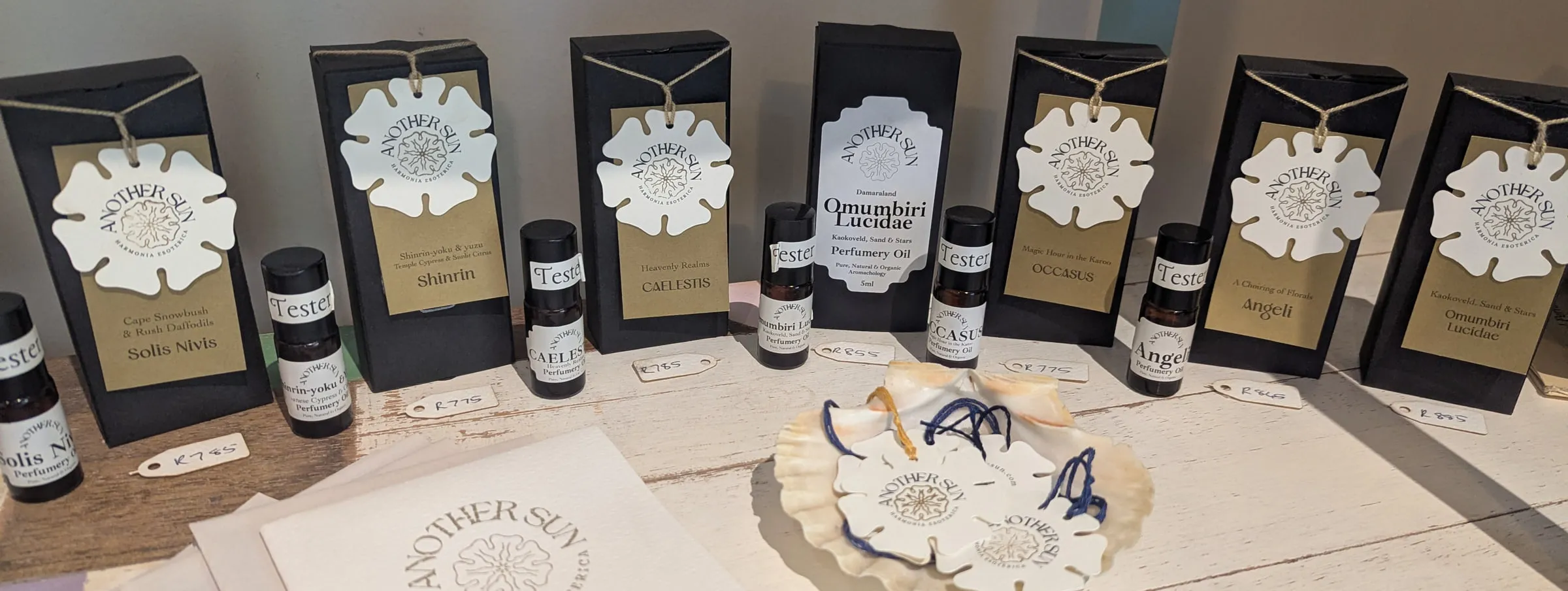 A row of Another Sun perfume oil boxes with white flower-shaped tags and small tester bottles