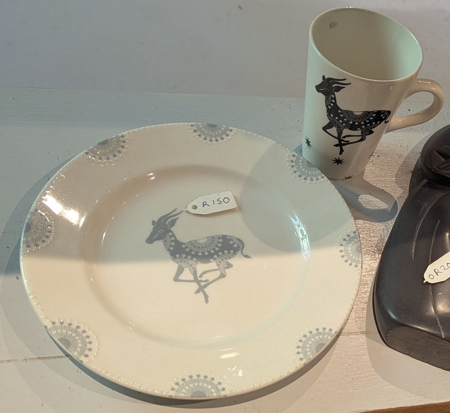 A white plate printed with a springbok and grey rosettes, with a matching mug behind