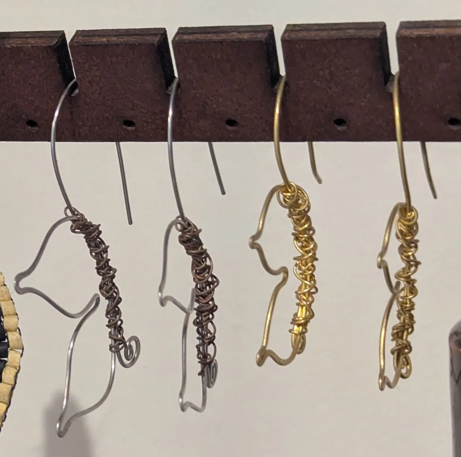 Four wire earrings shaped like birds, two silver-coloured with copper wrapping and two in brass, on a wooden earring bar