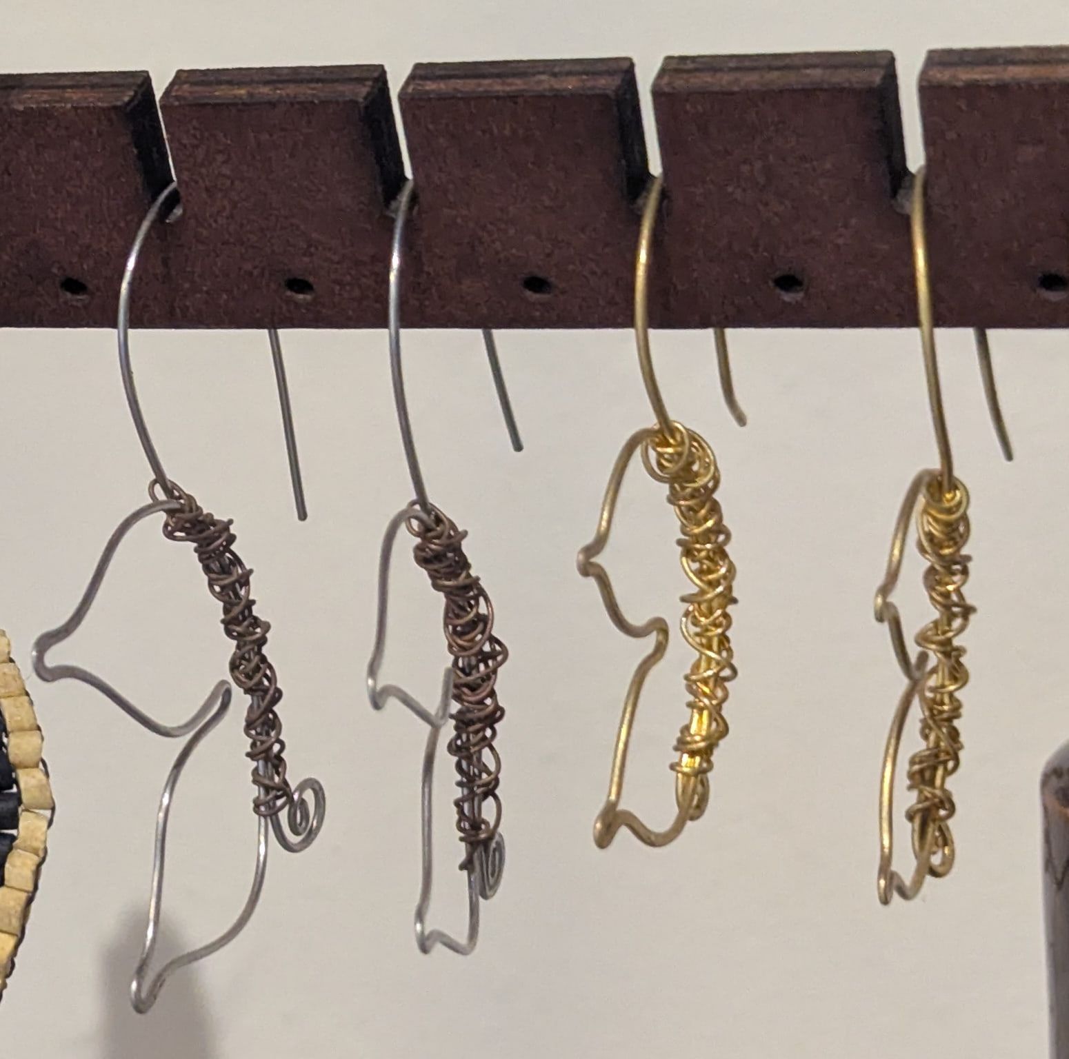Four wire earrings shaped like birds, two silver-coloured with copper wrapping and two in brass, on a wooden earring bar